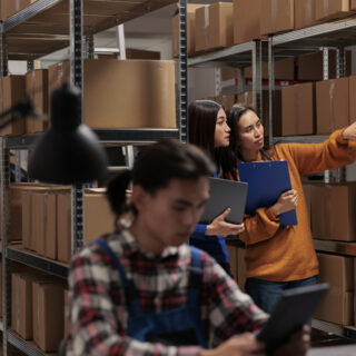 Storehouse asian women employees checking package packing quality in postal storage room. Storehouse shipment operator showing goods in stock on shelf to logistics manager