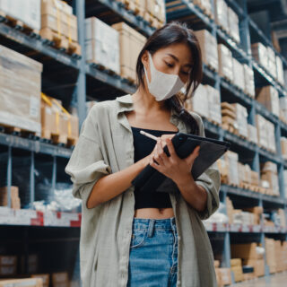 Young Asia businesswoman manager wear face mask looking for goods using digital tablet checking inventory levels stand in retail shopping center. Distribution, Logistics, Packages ready for shipment.
